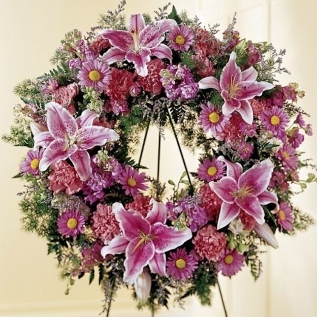 Standing funeral wreath of pink stargazer lilies, carnations, and mums with lush greenery on an easel.