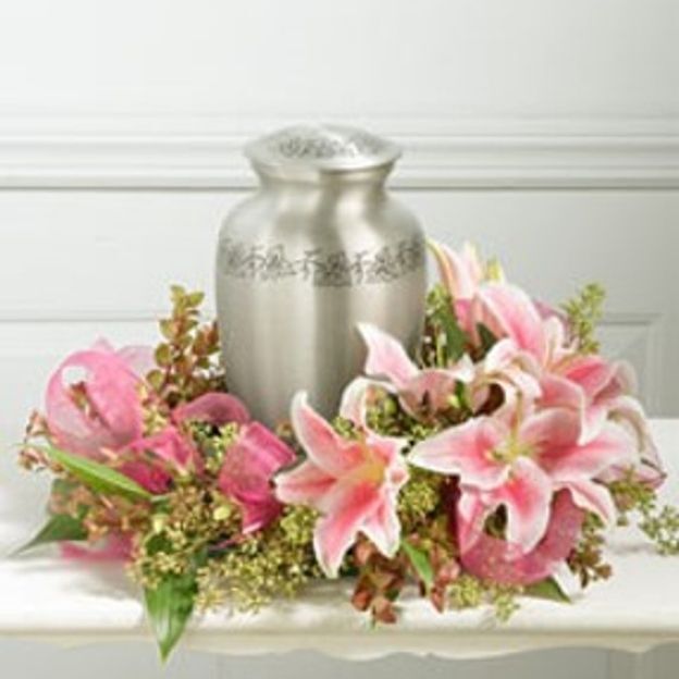 Silver memorial urn encircled by pink stargazer lilies and greenery on a white table.
