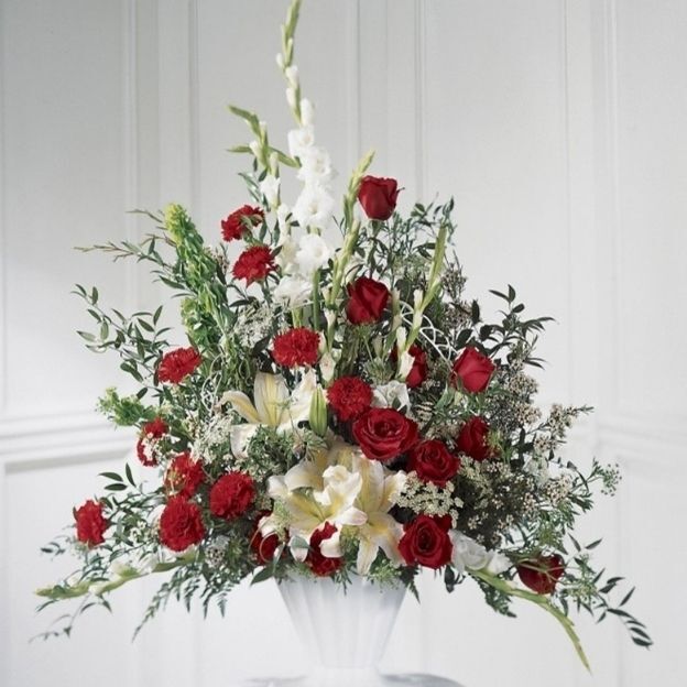 Sympathy arrangement of red roses and carnations with white lilies and gladiolus in a white pedestal vase.