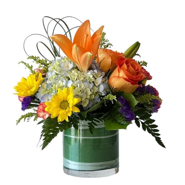 Bright mixed bouquet: orange lilies, orange roses, blue hydrangea, yellow mums, and greenery arranged in a short glass vase.