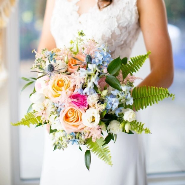 Close-up of wedding bouquet with soft pink and peach roses, blue flowers, and greenery held by bride in white dress.