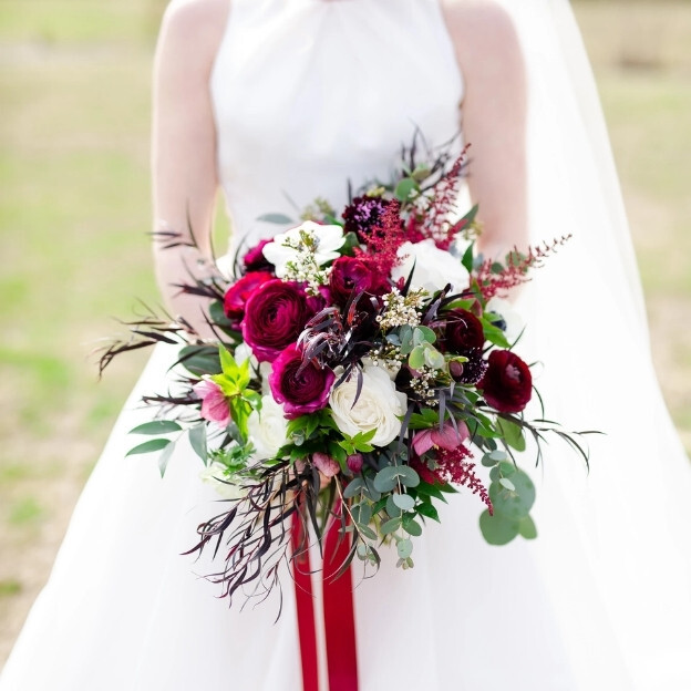 Elegant wedding bouquet featuring deep red and white roses, greenery, and textured accents arranged for a bridal portrait outdoors.