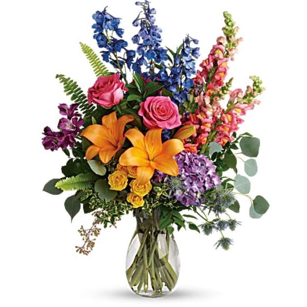 Vibrant arrangement of orange lilies, pink roses, blue delphinium, purple hydrangea, and eucalyptus in a clear vase.