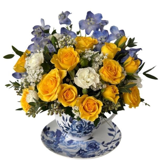 Compact bouquet of yellow roses, white carnations, and blue delphinium in a porcelain teacup with blue floral pattern.