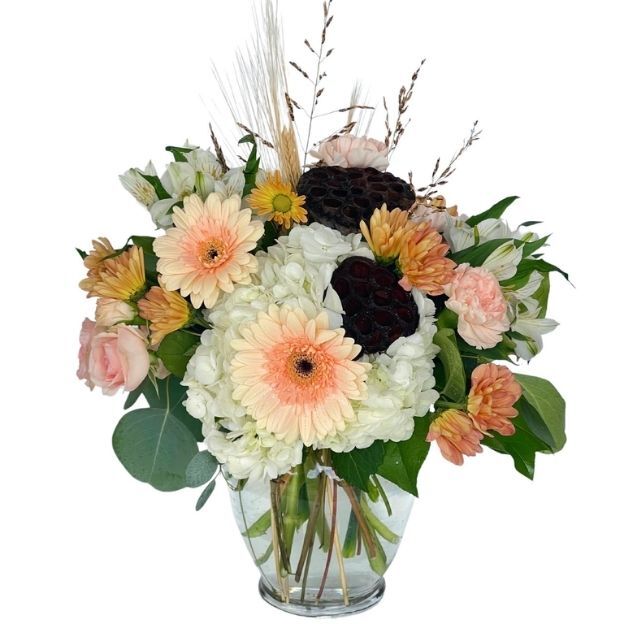 Soft autumn mix of peach gerberas, white hydrangea, mums, eucalyptus, and lotus pods with wheat in a clear vase.